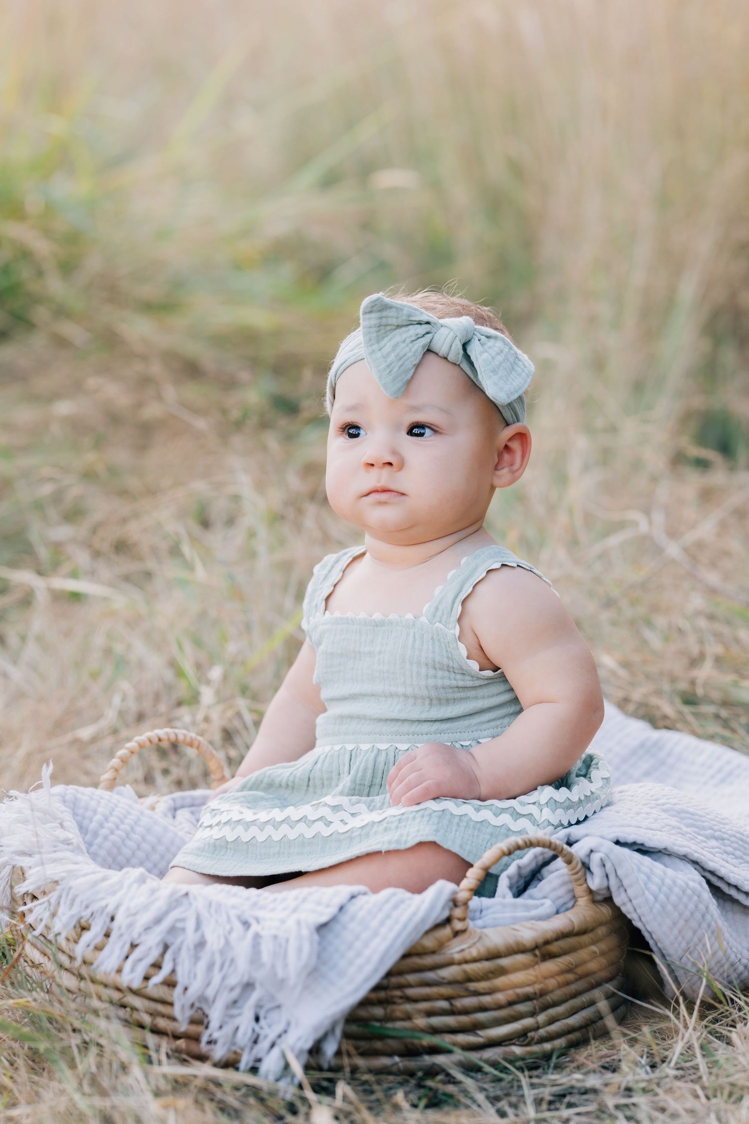 Adorable baby girl in sage green dress and bow headband sitting in wicker basket outdoors in golden grass field.