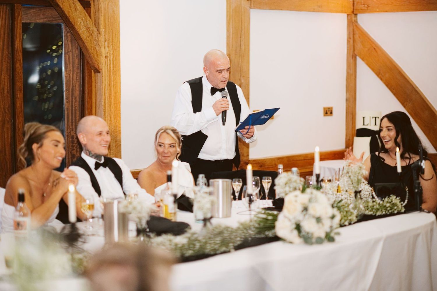 Man in a bow tie giving a speech as the top table smiles, Sandhole Oak Barn wedding