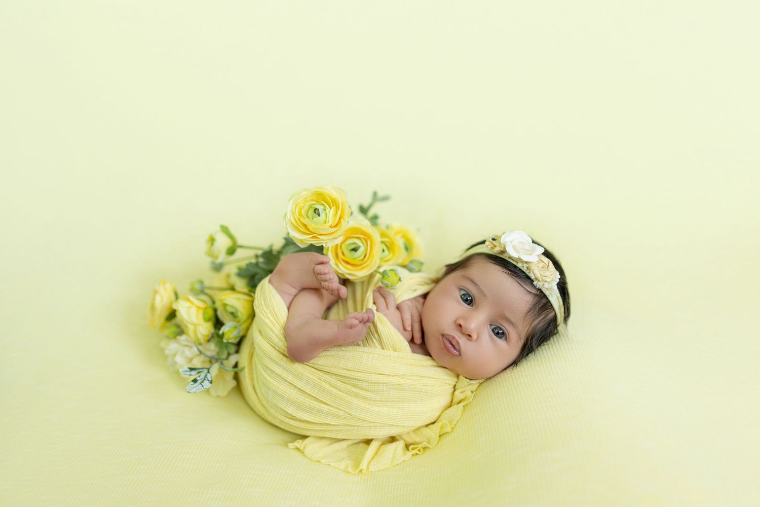 Newborn baby wrapped in yellow swaddle with floral headband surrounded by yellow roses on soft green background.