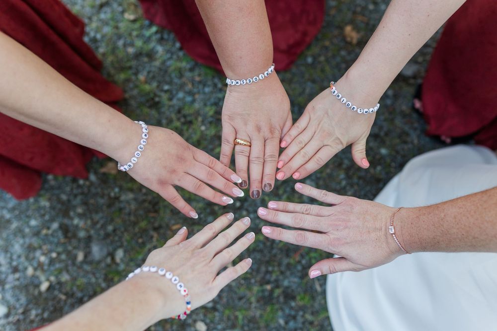 Bridesmaids in burgundy dresses and bracelets reach their hands together in a circle with a bride in white.