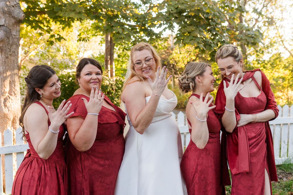 Bride in a white gown and four bridesmaids in red dresses smile and show off their rings outdoors by a white fence.