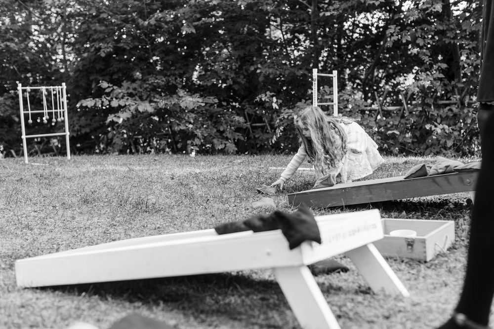 Woman in a long-sleeved top leans over a wooden cornhole board on a grassy lawn, with ladder toss games and trees in the background.