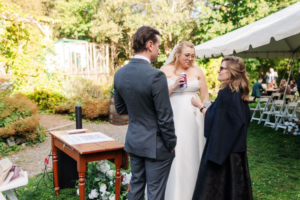 Bride in a white gown and groom in a gray suit stand beside an officiant at an outdoor garden wedding ceremony with a signing table.