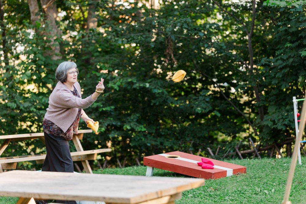Gray-haired woman in a pink cardigan tosses a bean bag toward a red cornhole board on a sunny lawn surrounded by green trees.