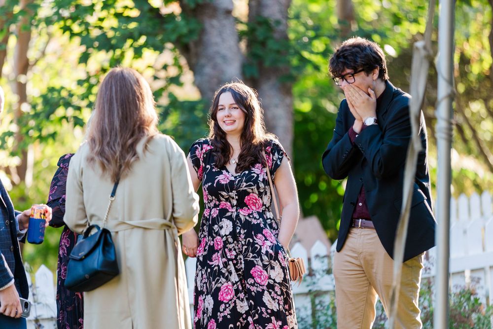 Smiling woman in a floral dress laughs with friends outdoors while a man in glasses covers his face in amusement.