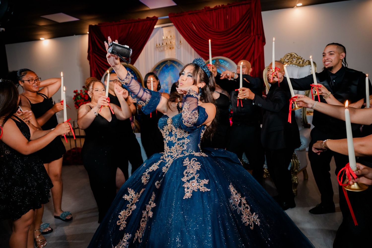 Quinceañera in a sparkling blue embellished ball gown takes a photo as guests hold lit candles at an elegant celebration with red drapes.