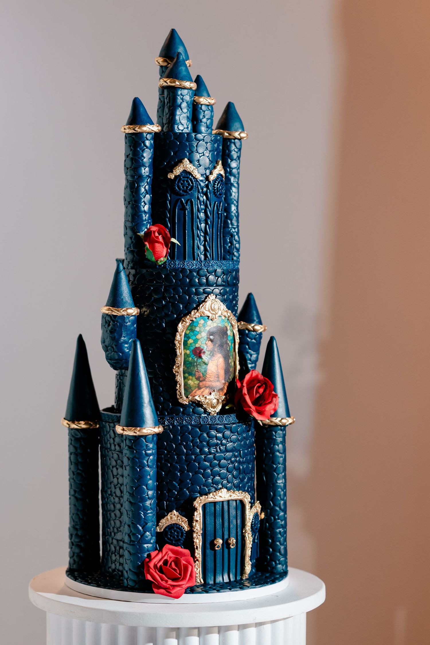 Tall navy blue castle wedding cake with gold-trimmed turrets, red sugar roses, and a framed fairytale portrait on a white pedestal.