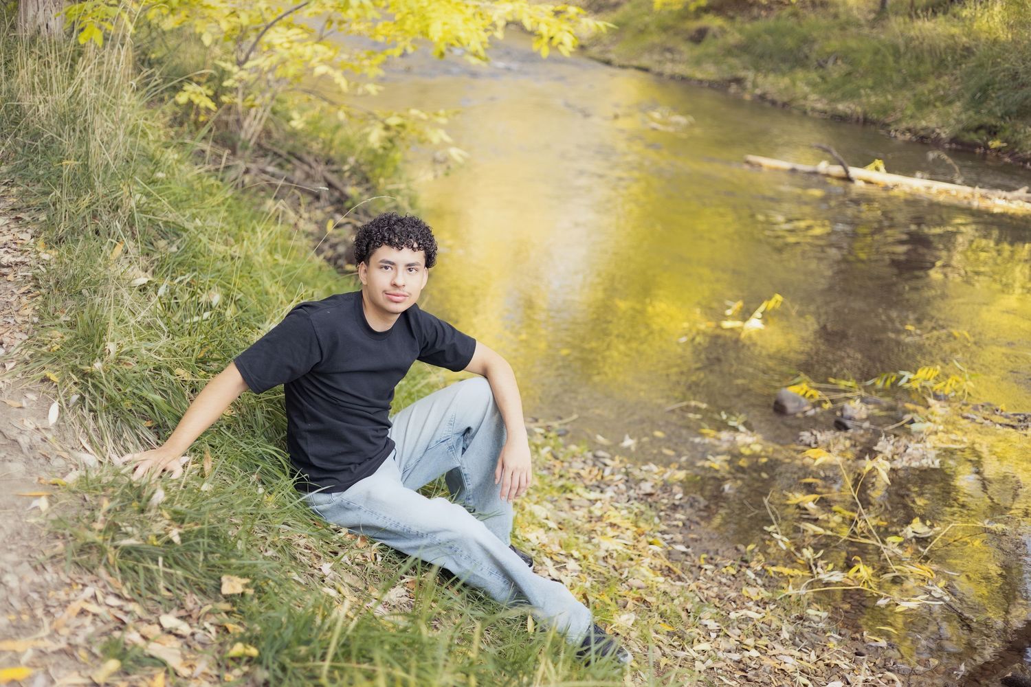 Young man in black shirt and jeans sitting on grassy riverbank surrounded by autumn foliage.