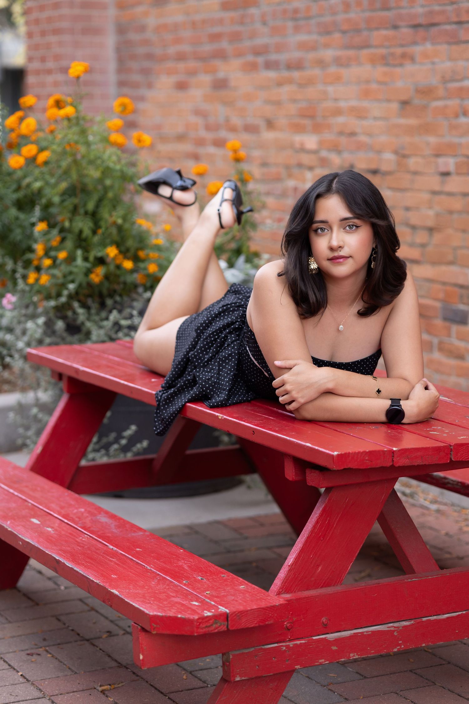 Young woman in black polka dot dress lying on a red picnic table outdoors with orange flowers and brick wall behind her.