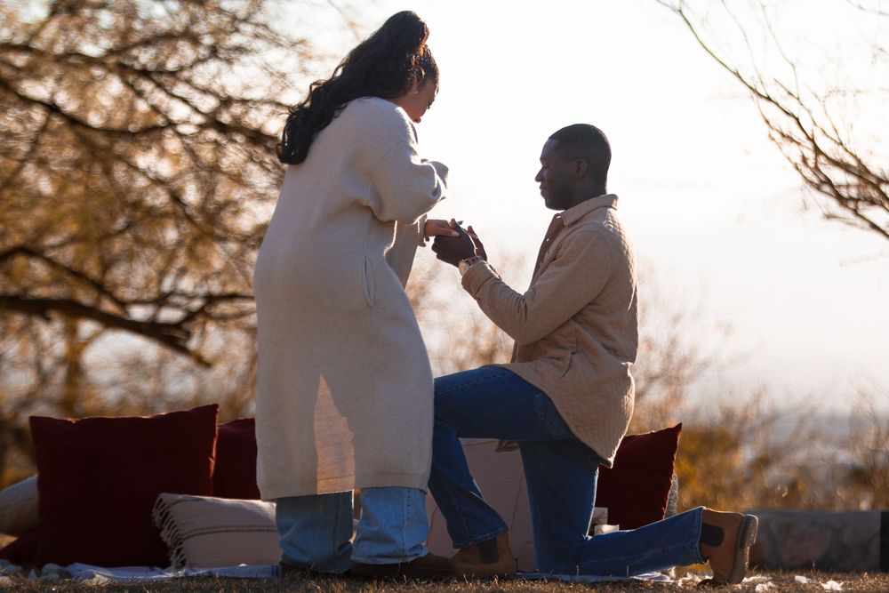 Man proposing to woman outdoors at sunset, kneeling on one knee with ring, romantic setting with cushions and bare trees.
