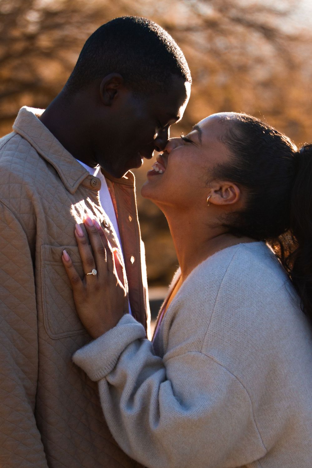 A romantic Black couple sharing a tender kiss outdoors in warm golden sunset light, woman wearing an engagement ring.