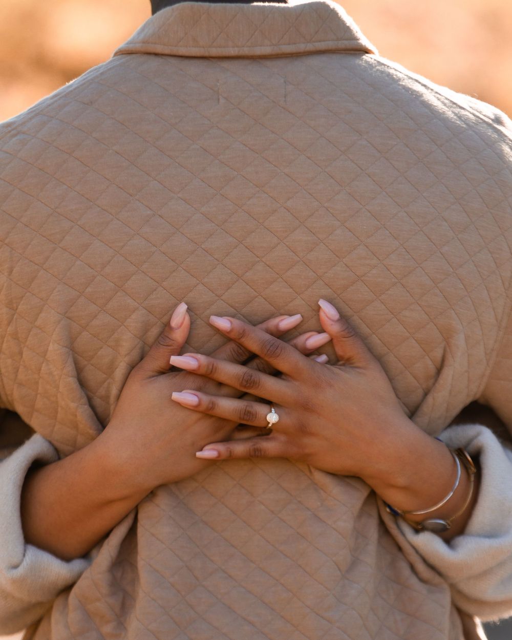 Person wearing a beige quilted jacket with hands crossed behind their back, showcasing pink manicured nails and a delicate bracelet.