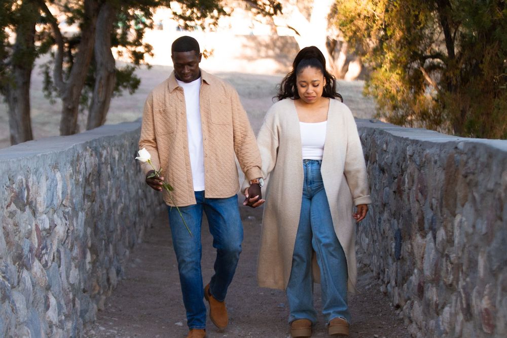 Couple holding hands walking on a stone bridge path outdoors, dressed in casual beige and denim outfits.