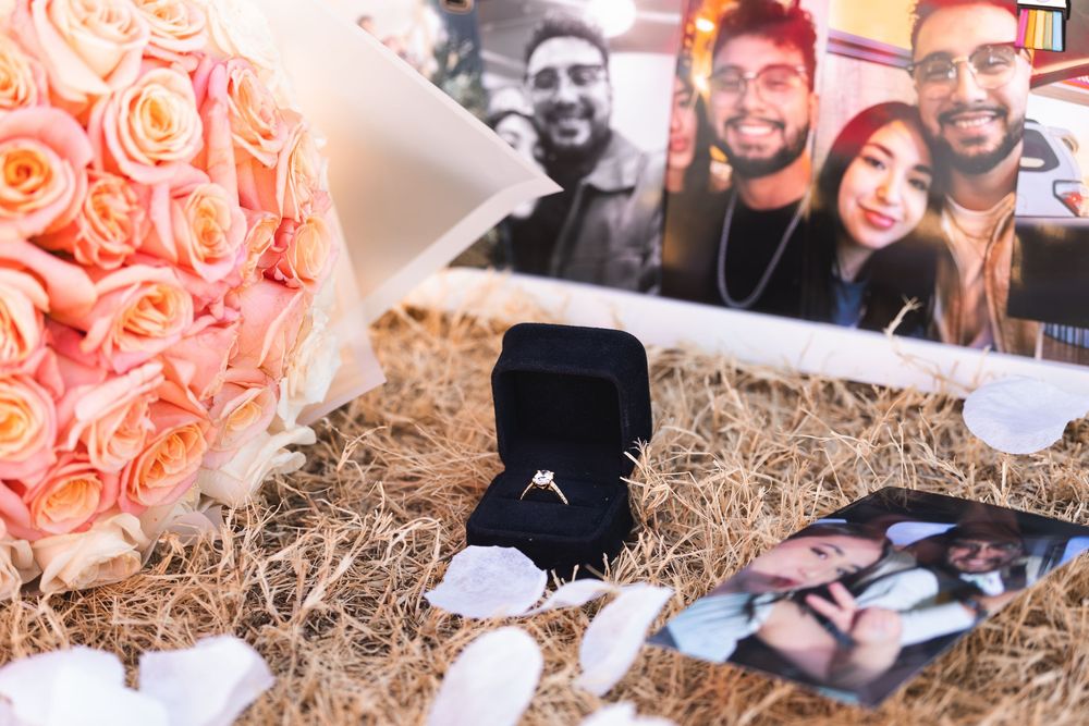 Engagement ring in black velvet box surrounded by pink roses, photos, and straw in a romantic proposal setup.
