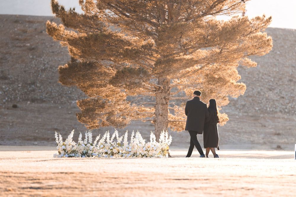 Couple walking hand in hand near a large golden tree and white floral arrangement in a serene desert landscape.