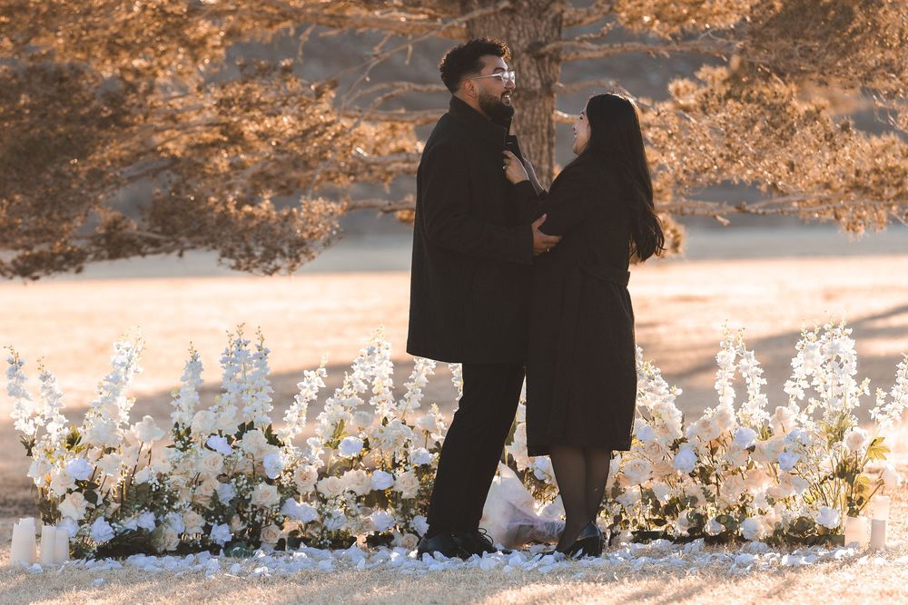 Couple in black outfits standing together near white floral arrangement outdoors during golden hour.