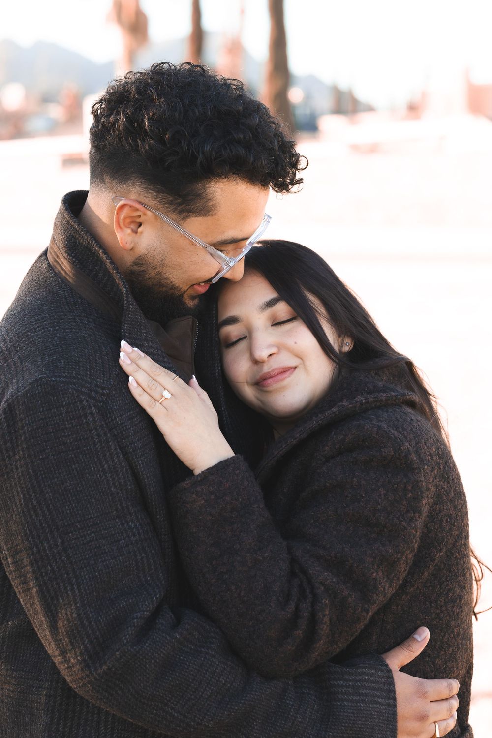 A couple in matching dark coats embrace tenderly outdoors, the woman smiling with eyes closed as the man leans in close.