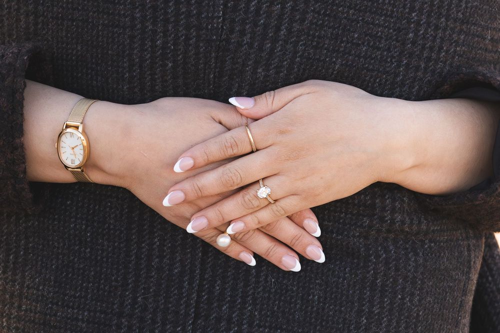 Close-up of woman's hands with French tip nails, gold watch, and delicate gold rings on dark background.