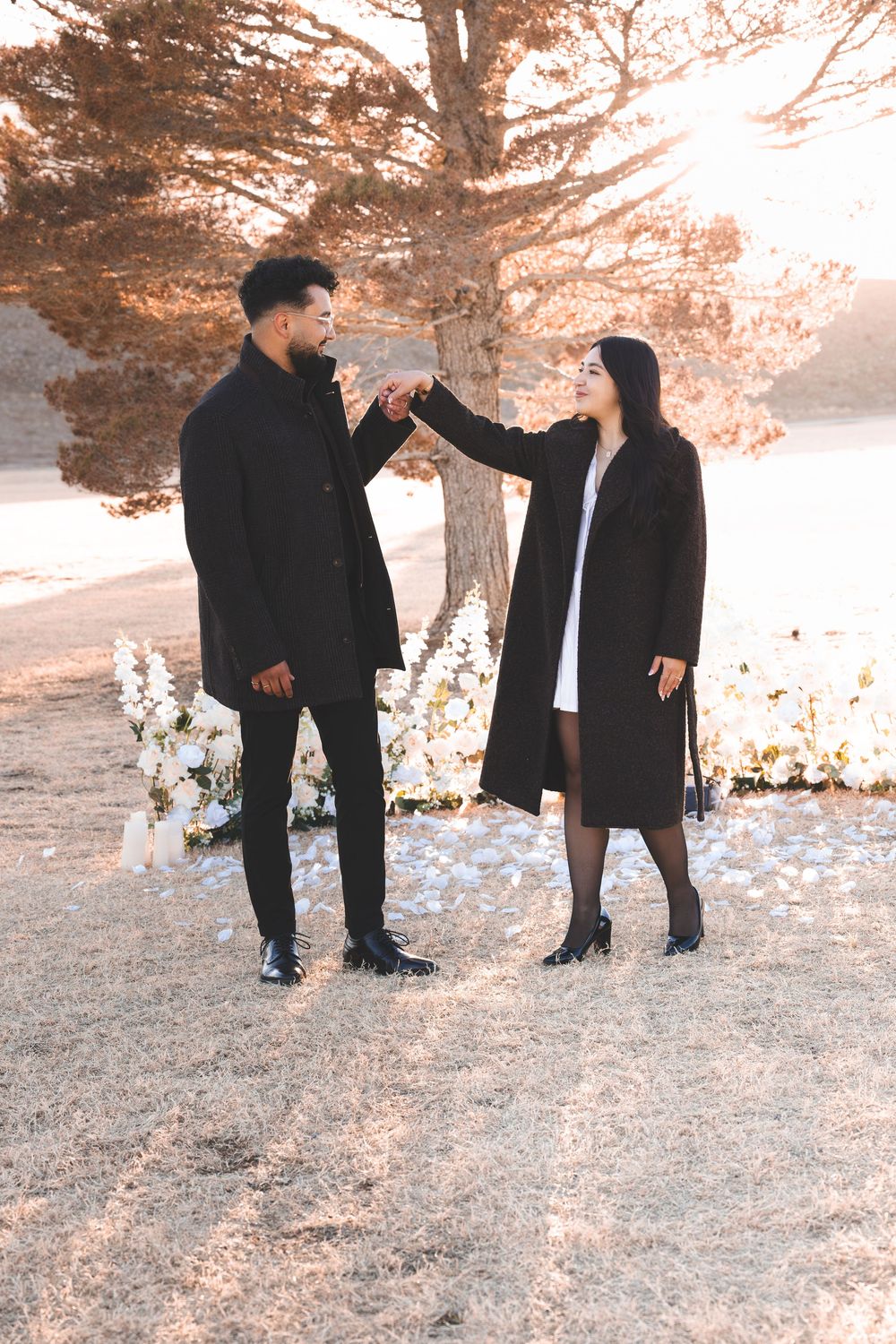 Couple in matching black coats holding hands outdoors near white floral arrangement and autumn tree in warm golden light.