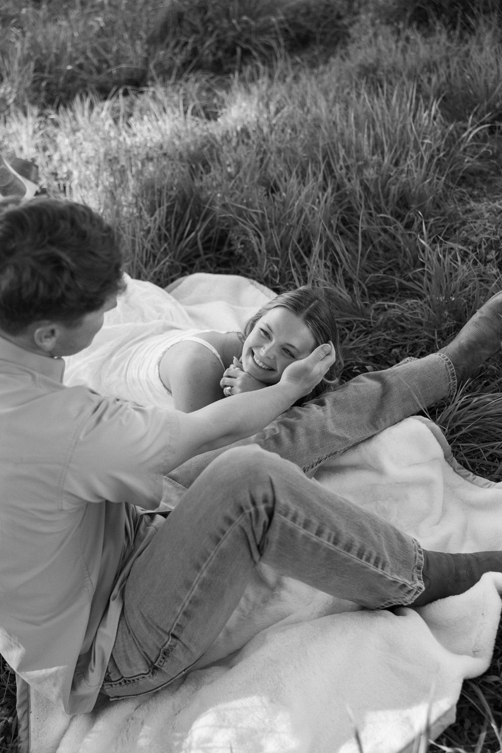 Black and white photo of a couple relaxing on a blanket in a field, woman smiling while lying down.