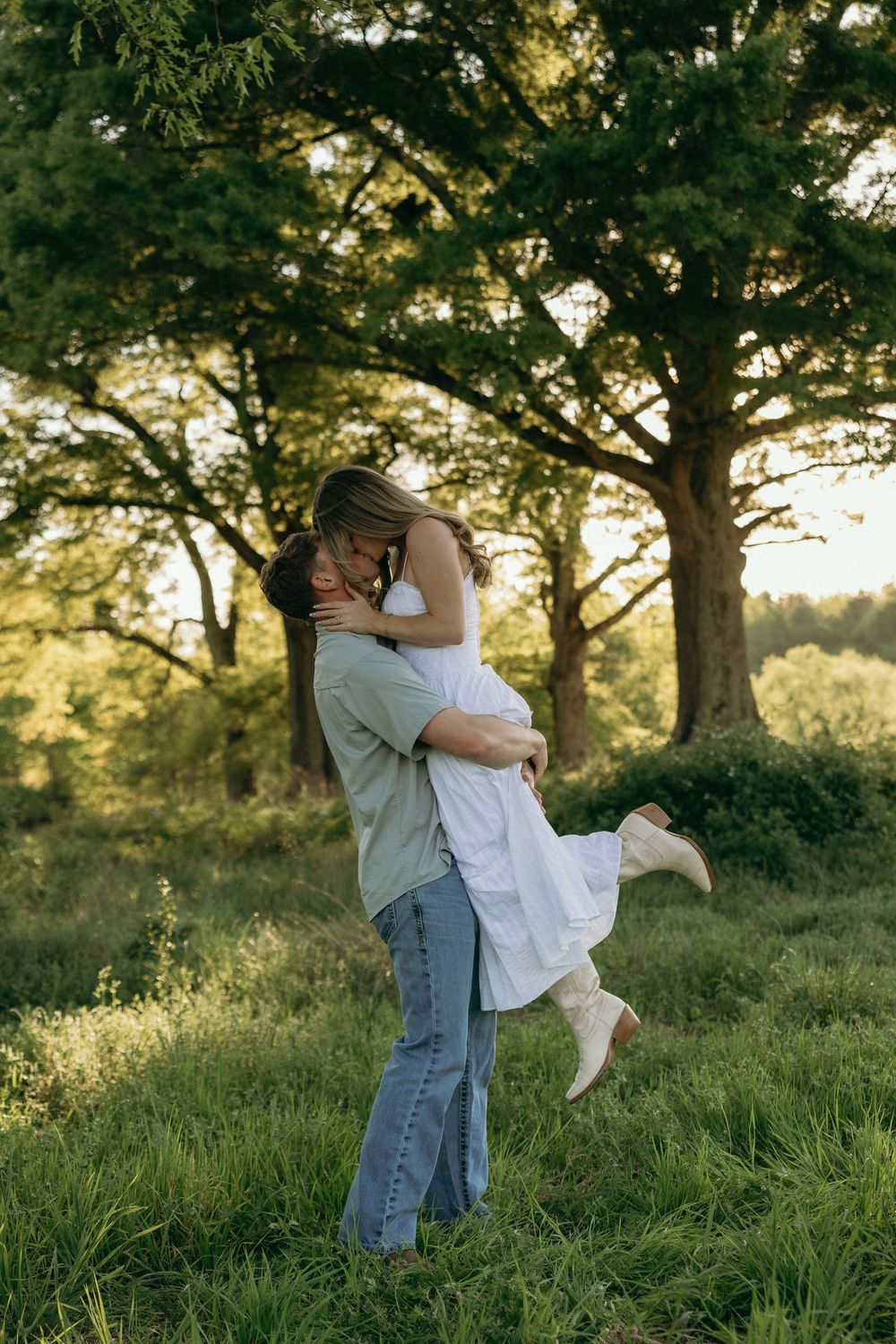 A couple shares a romantic moment as a man lifts a woman in a white dress outdoors in a lush green field.