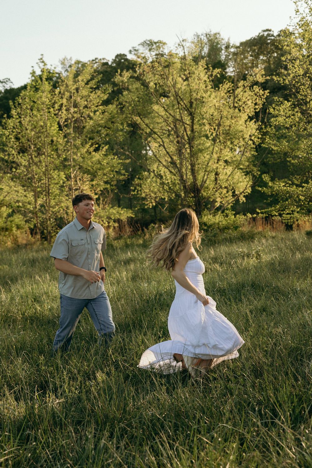 A couple dances playfully in a sunlit green meadow, the woman in a white dress twirling as the man smiles at her.