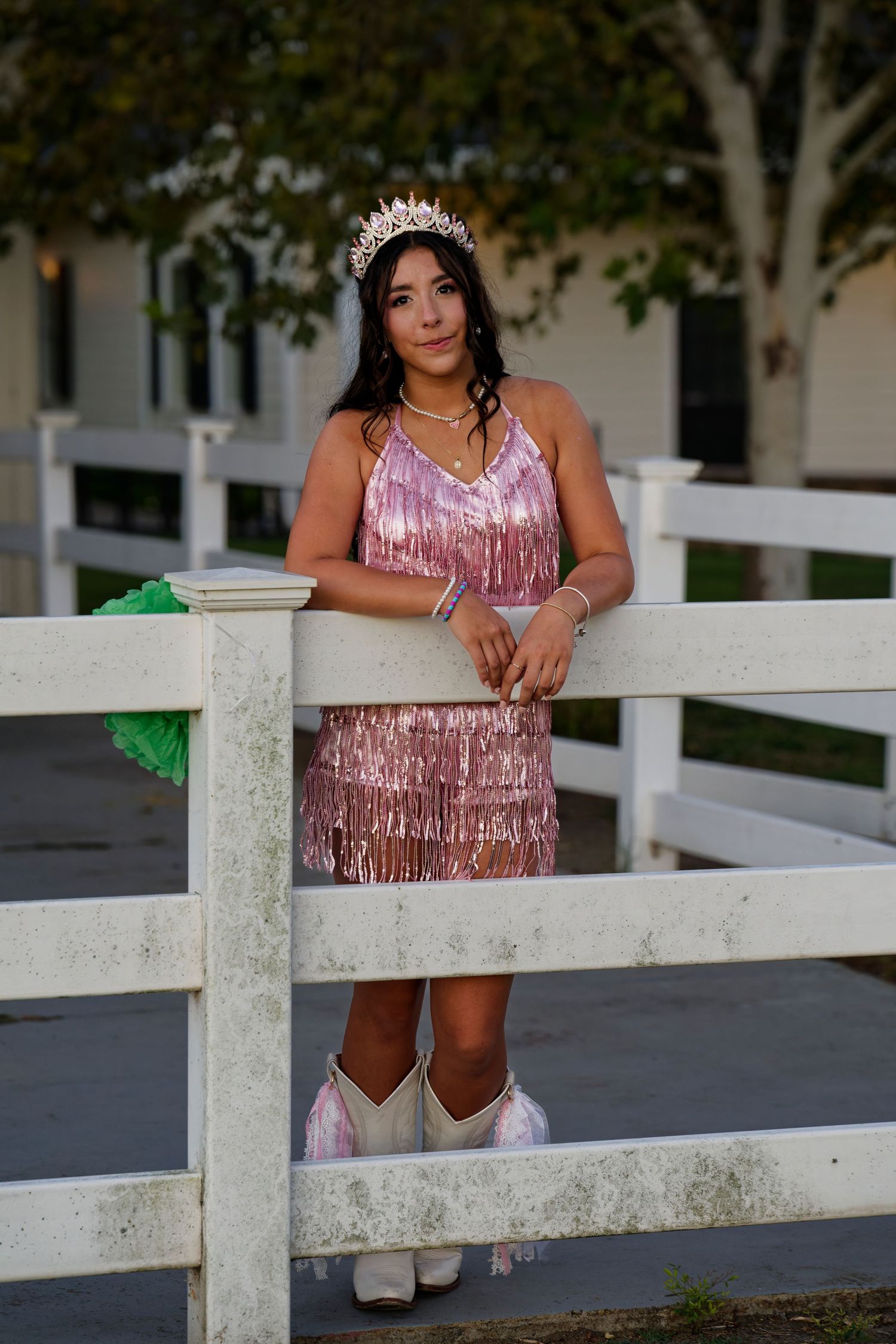 A quinceañera girl in a pink sequined dress and white cowboy boots poses by a white fence outdoors.