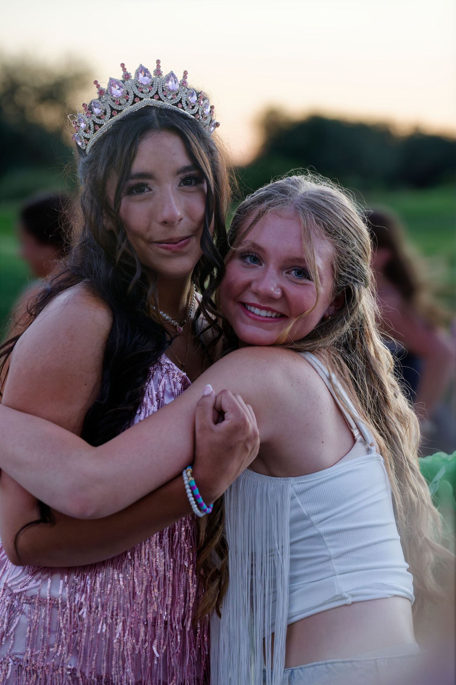 A quinceañera girl wearing a tiara in a pink fringe dress hugs a smiling friend at her outdoor celebration.