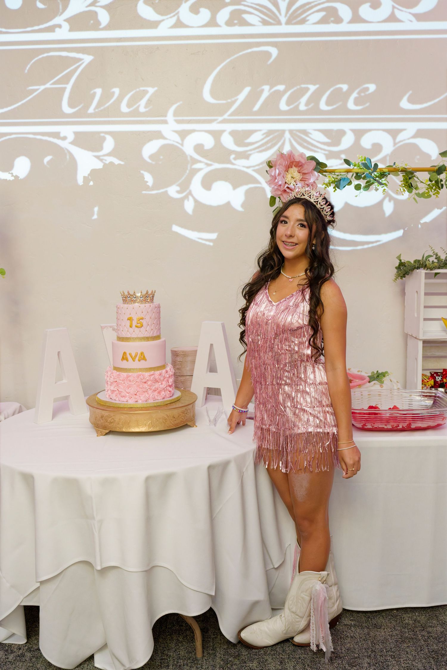 Young woman in pink fringe dress and tiara stands beside quinceañera cake at Ava Grace celebration party.