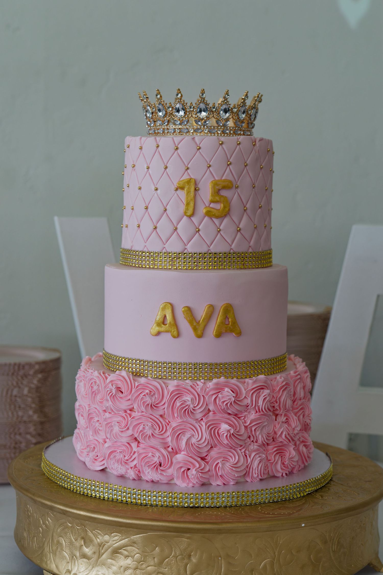 Three-tier pink and gold quinceañera cake with crown topper reading 15 and AVA on golden stand.
