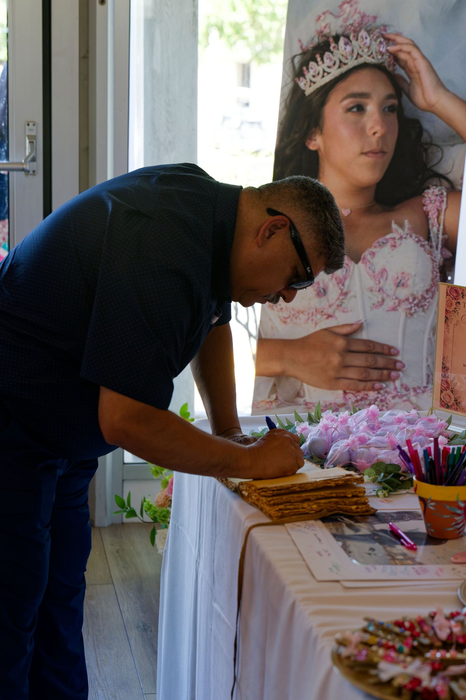 Guest signs guestbook at quinceañera welcome table beside large portrait of girl in tiara and pink gown.
