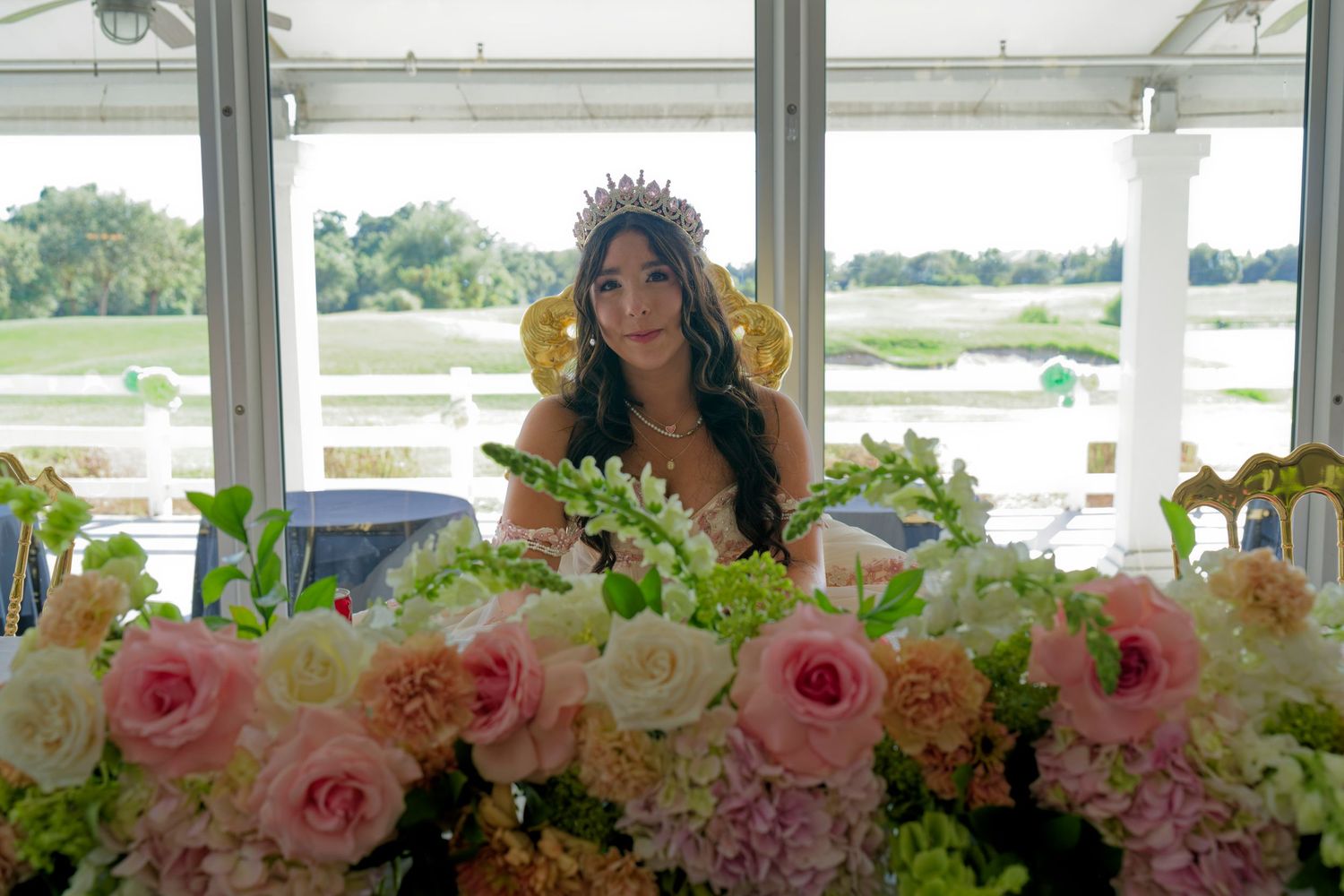 A quinceañera girl wearing a tiara sits behind an elegant floral arrangement of pink and white roses.