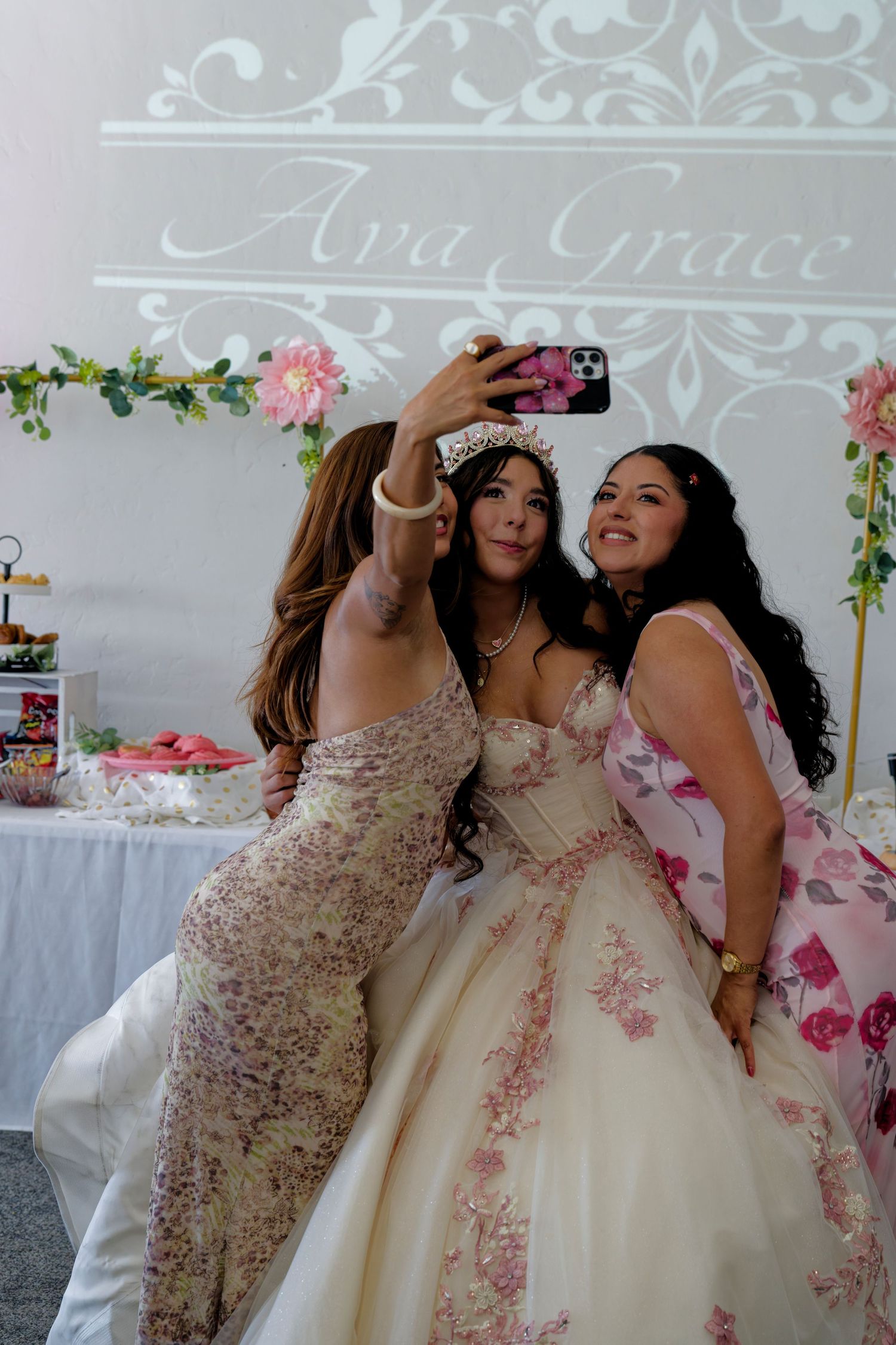 A quinceañera girl in a white floral gown takes a selfie with two guests at her Ava Grace celebration.