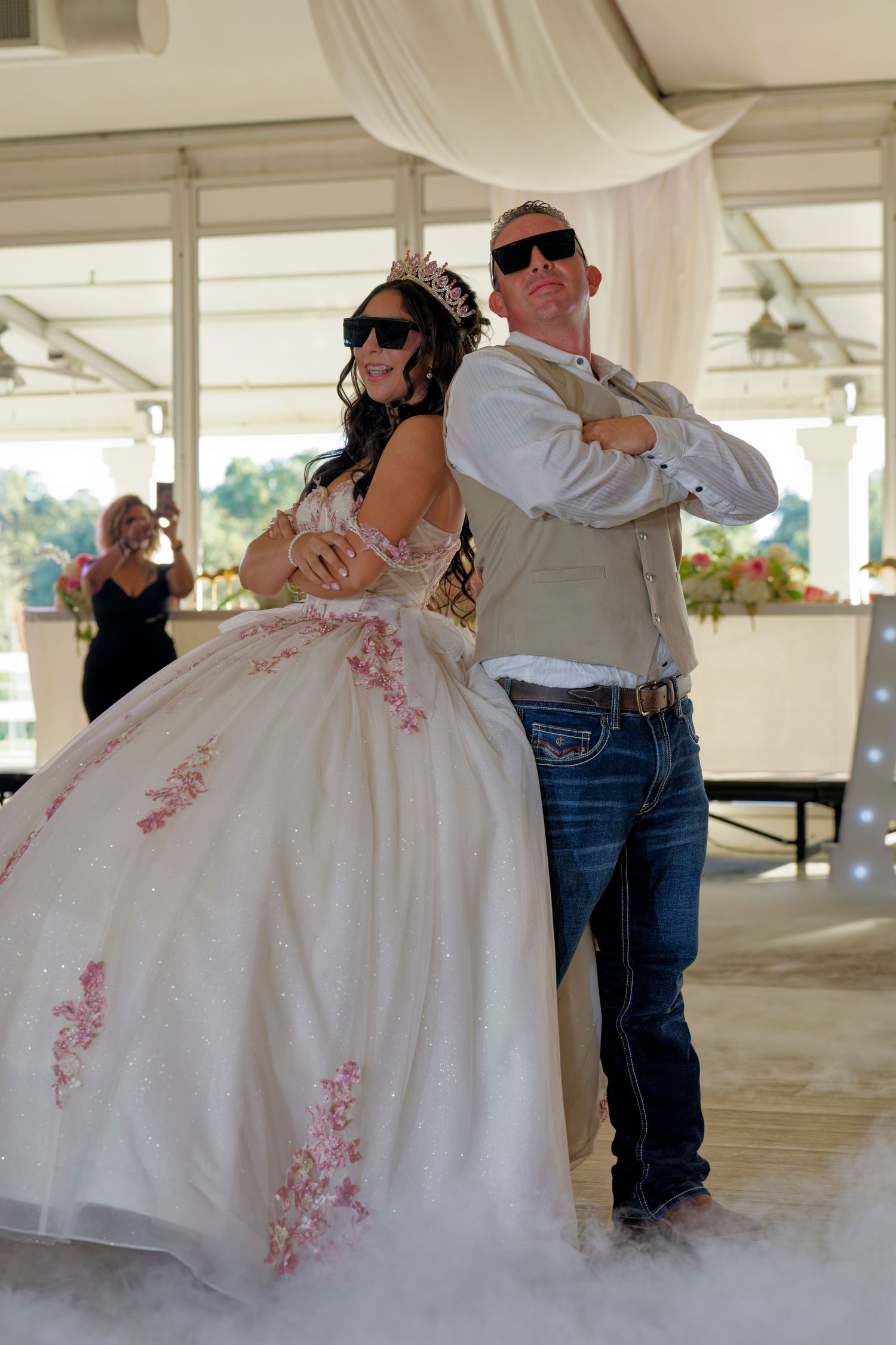 A quinceañera girl in a white and pink ballgown poses back-to-back with a man in sunglasses at her celebration.