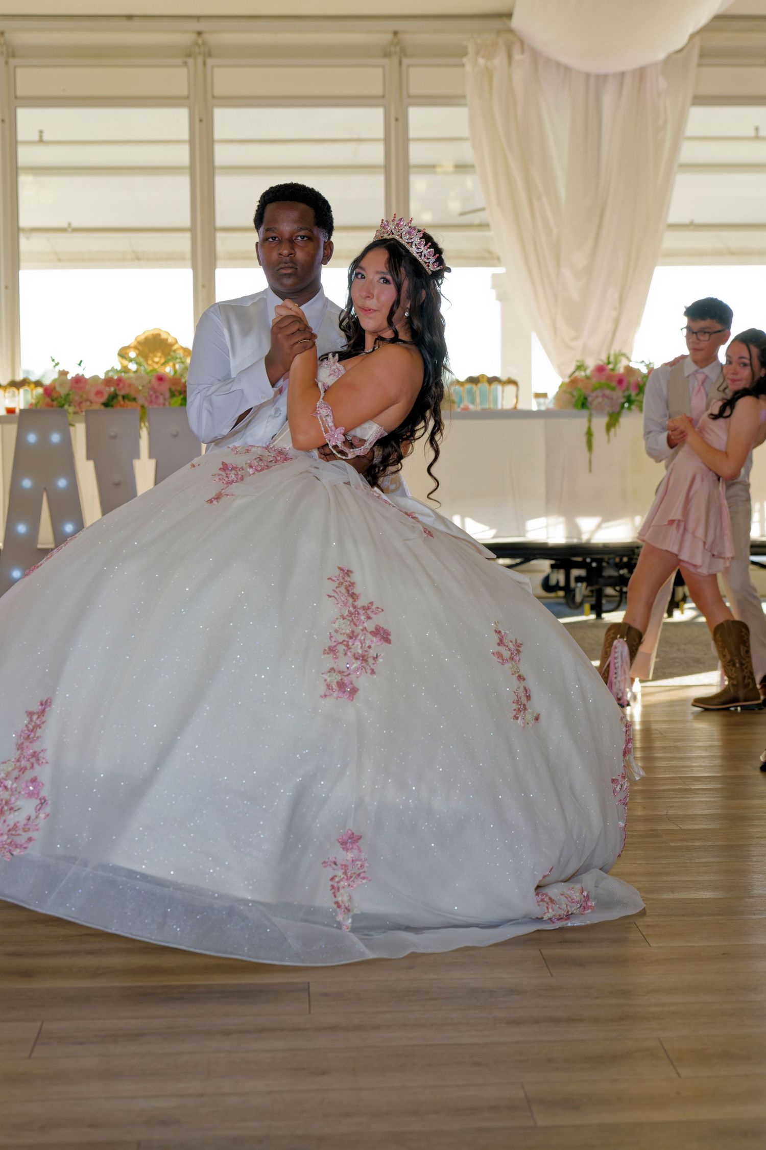 A quinceañera girl in a white pink floral ballgown shares a first dance with a young man at her reception.