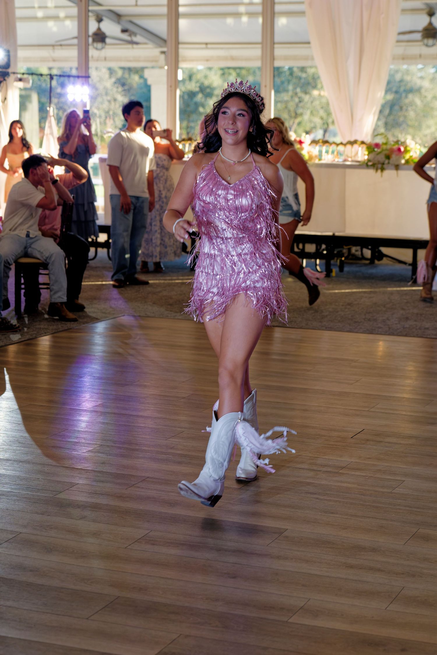 A quinceañera girl in a pink sequined fringe dress and white cowboy boots dances at her celebration.
