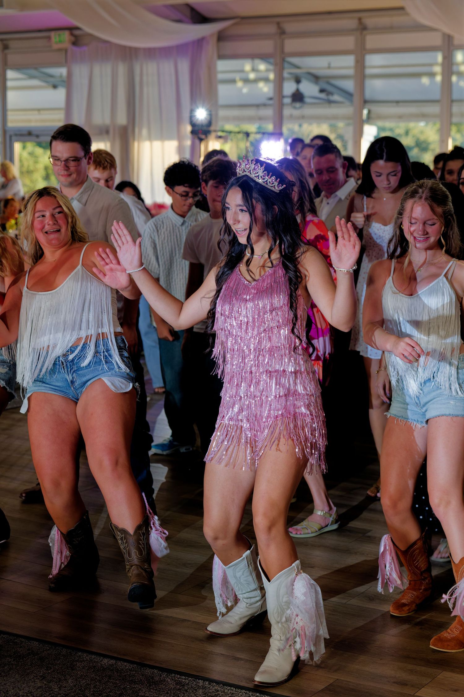 A quinceañera girl in a pink fringe dress and tiara dances with her court in matching outfits at her party.
