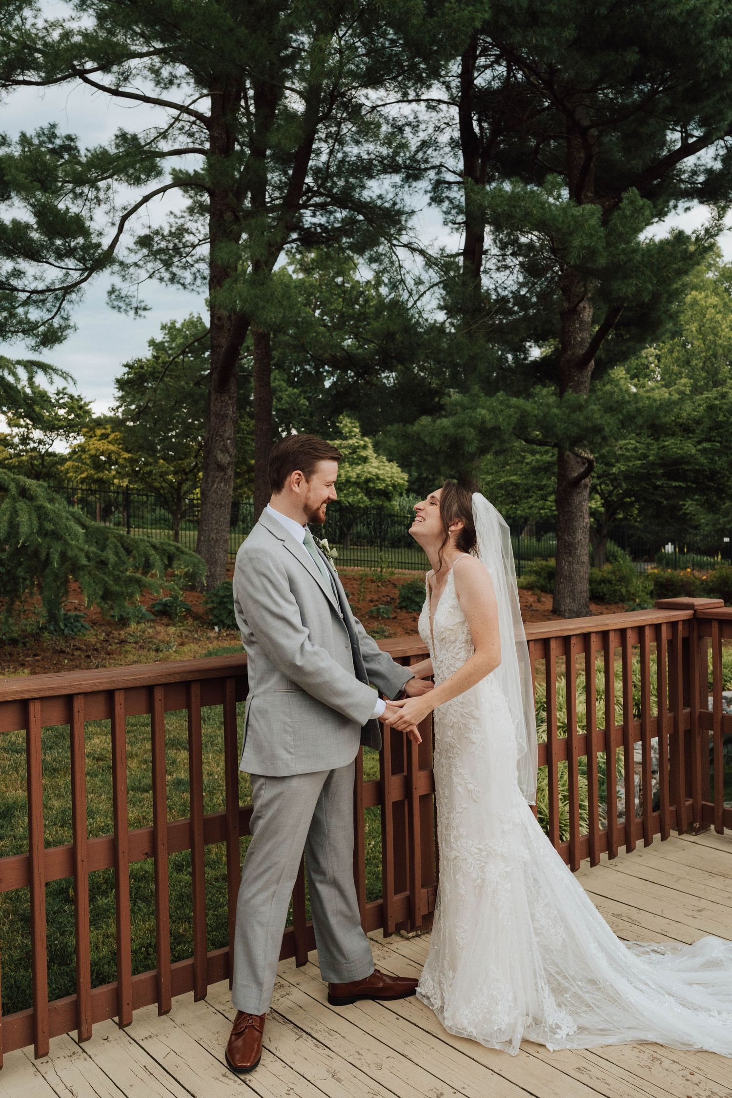 Bride in lace wedding dress and veil walks toward groom on wooden deck surrounded by lush green trees.