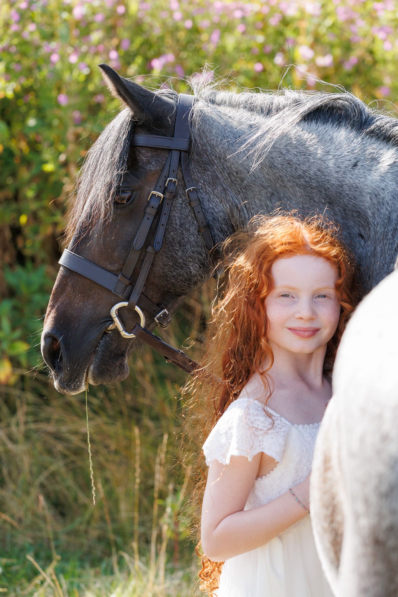 Young woman smiling between two bay horses with white markings, holding reins near a brick stable.