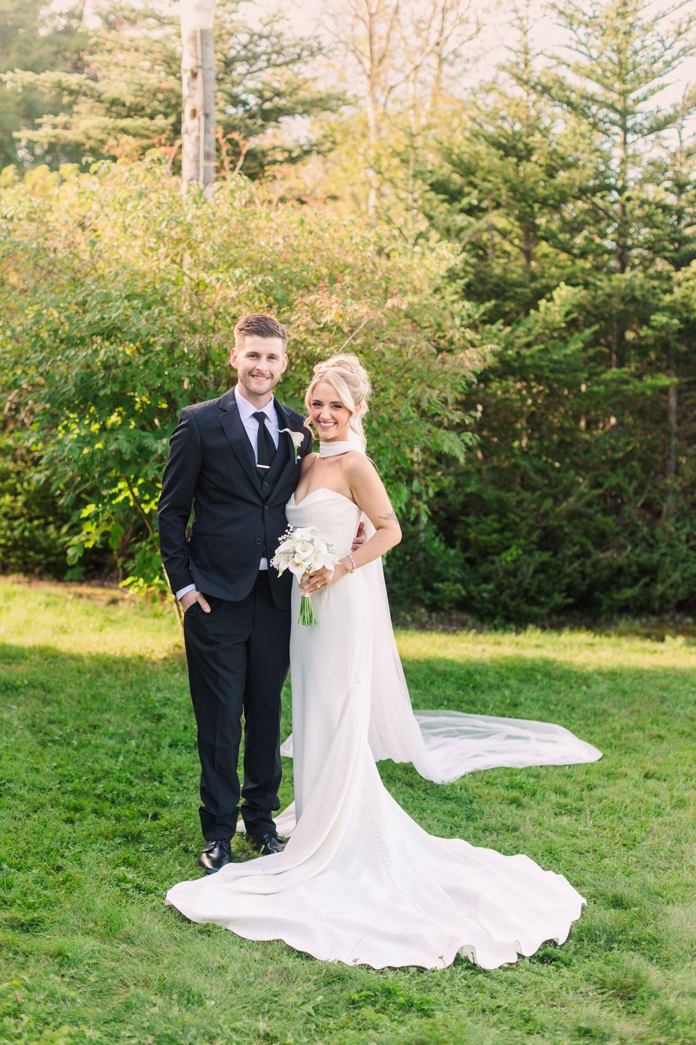 A bride in a white gown with a long train holds a bouquet beside a groom in a black suit on a green lawn.
