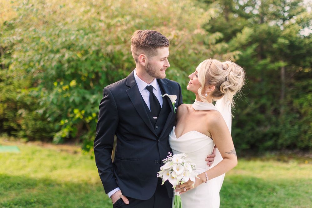 A bride and groom share a romantic moment outdoors, she holds a white bouquet in a strapless gown as he wears a navy suit.