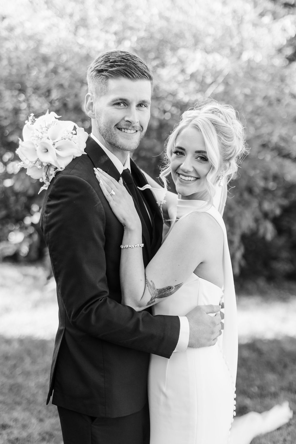 Black and white wedding portrait of smiling bride and groom embracing outdoors, bride holding bouquet.