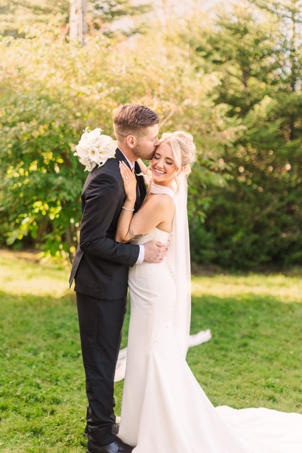 A bride and groom share a romantic embrace outdoors, with the groom kissing her cheek as she smiles holding white flowers.