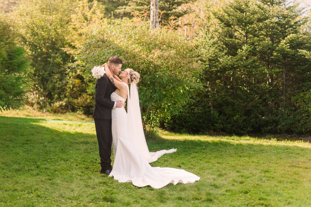 A bride and groom share a romantic embrace on a green lawn surrounded by lush trees on their wedding day.