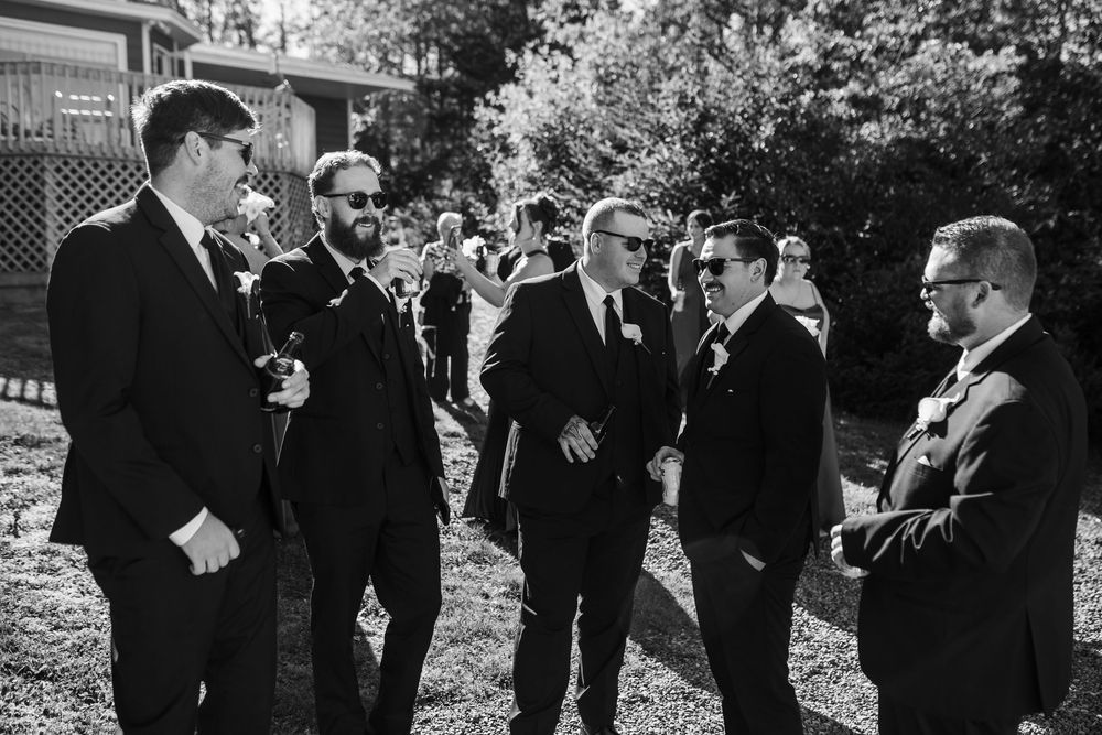 Groomsmen in black suits socializing outdoors at a wedding ceremony in a candid black and white photo.