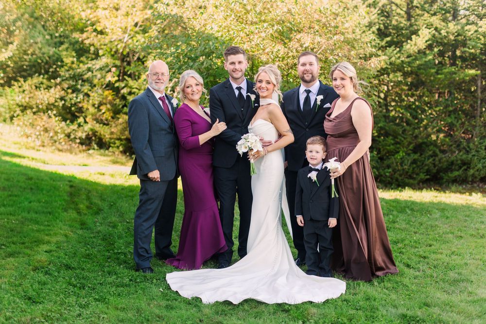 Wedding party poses outdoors on green lawn with trees, bride in white gown, groom in black suit, family in formal attire.