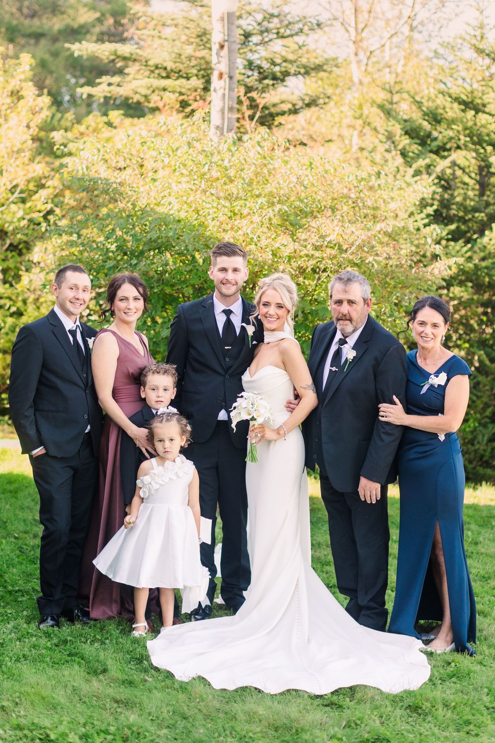 Wedding party poses outdoors on green lawn, bride in white gown with bouquet, groom in suit, family in formal attire, trees behind.