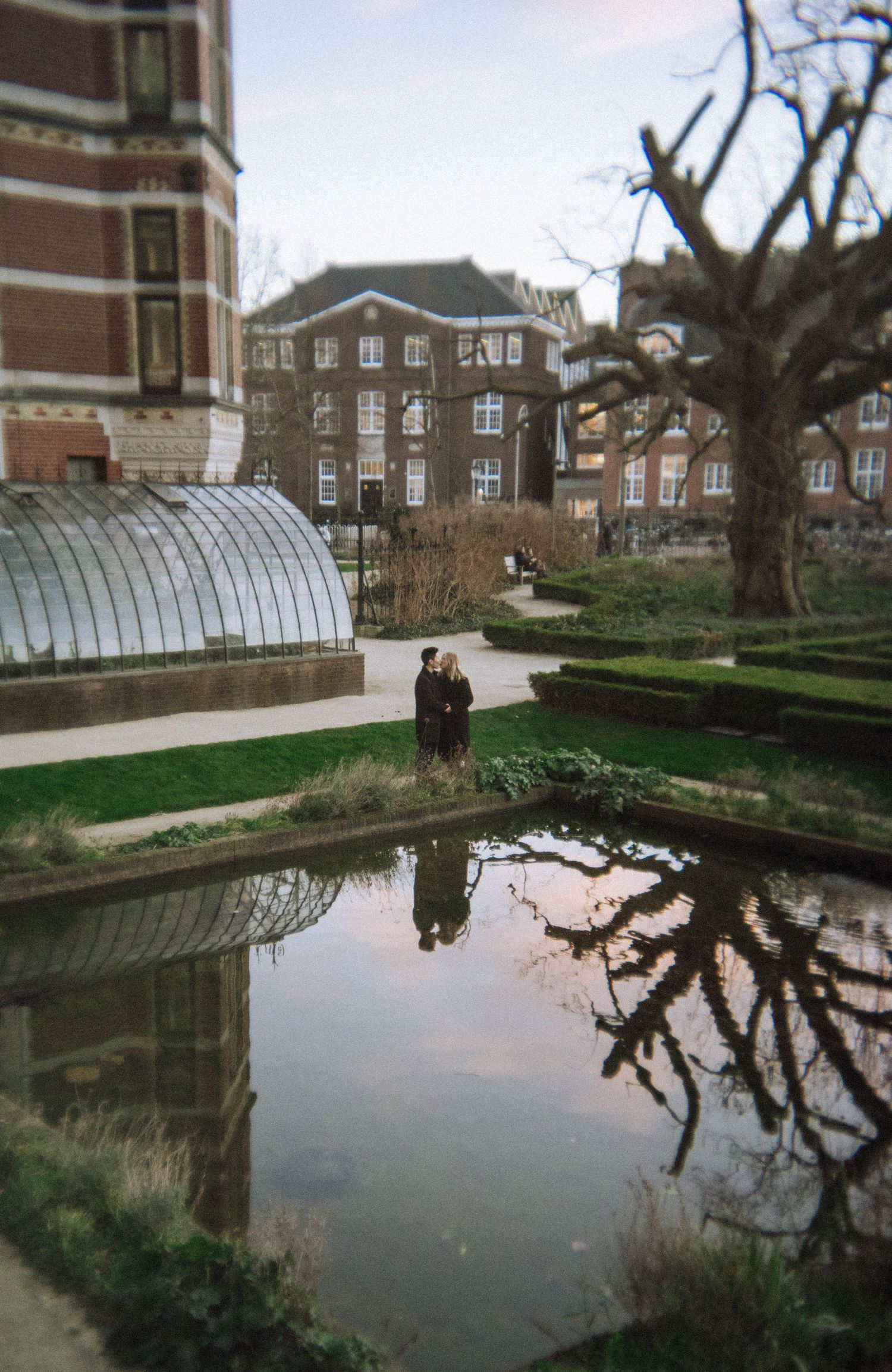 Two people stand together on a garden path beside a calm reflecting pond, with a glass greenhouse - Rijksmuseum gardens.
