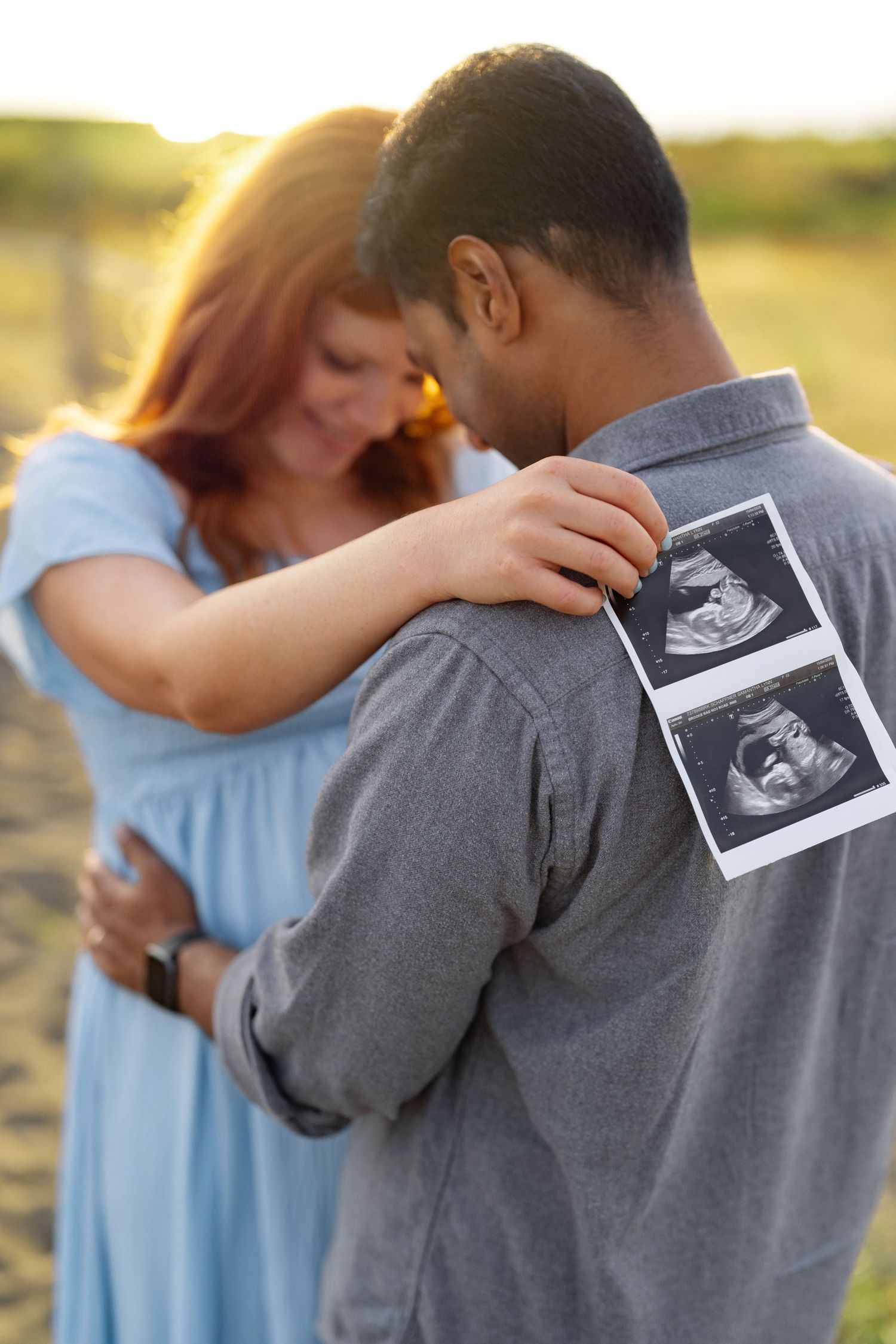 Expectant couple embracing outdoors at sunset, with two ultrasound photos pinned to the man's gray shirt in a pregnancy announcement.