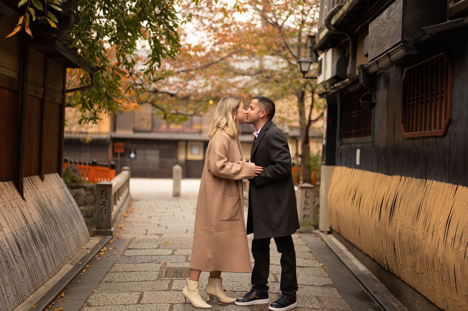 Couple in autumn coats share a kiss on a quiet stone-paved lane lined with traditional wooden buildings and fall foliage.